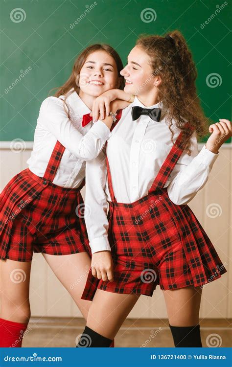 Two Brunette Schoolgirls In School Red Uniforms Are Standing In A Classroom With Books Stock