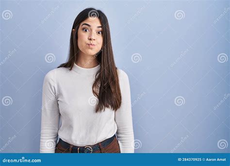 Young Brunette Woman Standing Over Blue Background Puffing Cheeks With Funny Face Stock Image