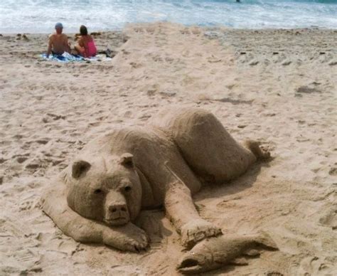 A Sand Sculpture Of A Bear Laying On The Beach With People In The Water