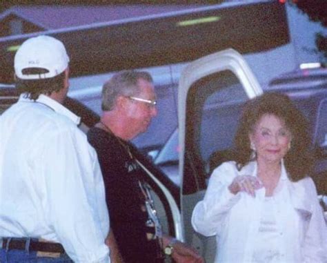 A Man And Woman Standing Next To Each Other In Front Of A White Car