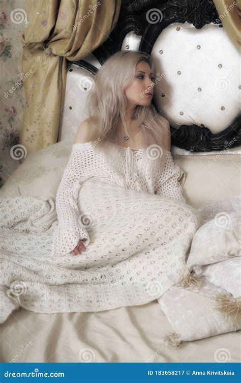 Beautiful Blonde With A White Knitted Sweater Sitting On The Bed Stock