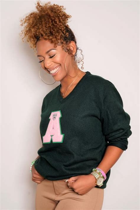 A Woman With Curly Hair Wearing A Green Sweater And Khaki Pants Smiles