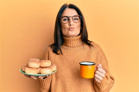 Middle Age Brunette Woman Eating Doughnuts And Drinking A Cup Of Coffee Looking At The Camera