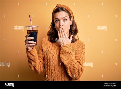 Une jeune fille blonde buvant un verre de soda sucrée sur fond jaune isolé couvre la bouche avec