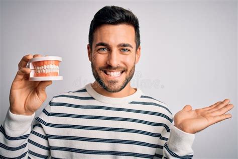 Young Handsome Man With Beard Holding Plastic Denture Teeth Over White