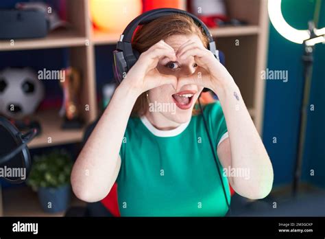 Young Redhead Woman Streamer Smiling Confident Doing Heart Symbol With Hands At Gaming Room