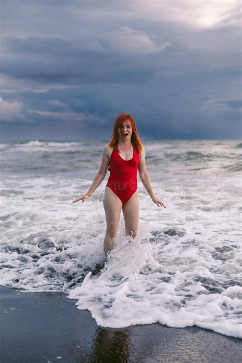 Happy Woman Tourist In Red Bodysuit Standing On Black Sand Big Stormy