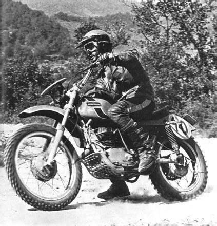 A Man Riding On The Back Of A Motorcycle Down A Dirt Road With Trees In