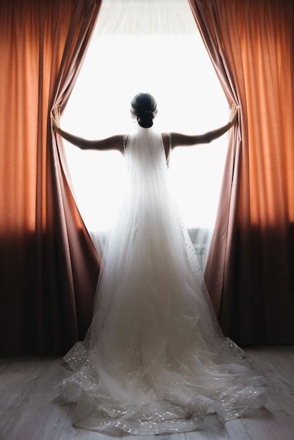 La novia y el novio están de pie contra la ventana mirando hacia afuera