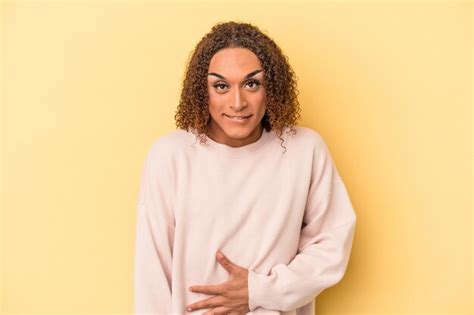 Joven transexual latina aislada de fondo amarillo toca la barriga sonríe suavemente come y el
