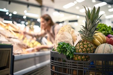 棚から製品を取り出している背景の女性の間に食品、果物、野菜でいっぱいのスーパーマーケットでのショッピングカートのクローズアップ 無料の写真