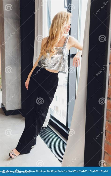 Beautiful Blonde Woman In Black Clothes With Heels Stands By The Window