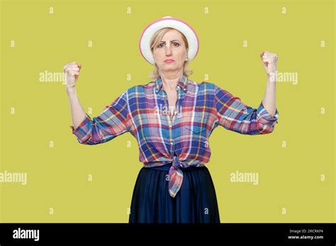 Portrait Of Mature Woman Wearing Checkered Shirt And Hat Raises Hands And Shows Muscles Feels