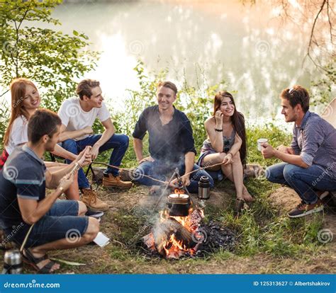 Group Of Friends Are Sitting Around Camp Fire And Preparing Sausages