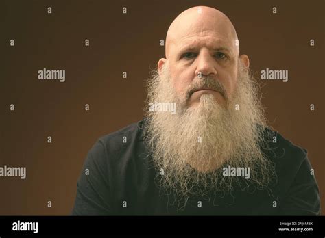 Studio Shot Of Mature Bald Man With Long Beard Sitting On Chair Against