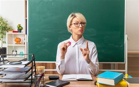 Jeune enseignante blonde sérieuse portant des lunettes assis au bureau avec des outils scolaires