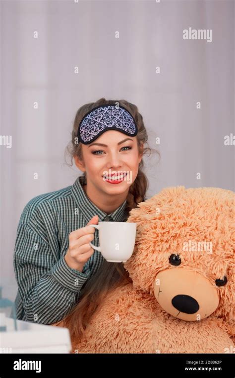 Young Woman Sitting With White Cup And Toy Bear Smiling In Sleep Mask