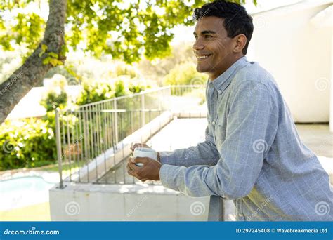 Happy Biracial Man Holding Mug And Standing On Balcony At Home Stock