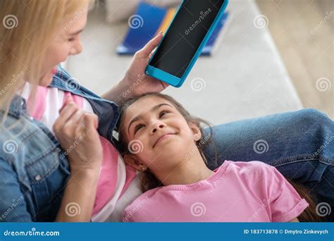 Blonde Female Siting On Sofa Holding Tablet Dark Haired Girl Lying On Her Lap Smiling Stock