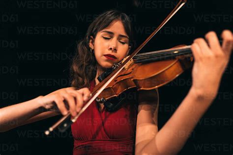 Woman In Red Dress Playing Violin With Closed Eyes During Concert On Stage In Dark Theater Stock