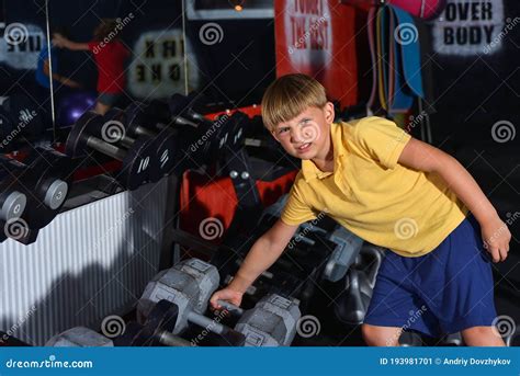 A Preschooler Boy Is Trying To Lift A Heavy And Large Dumbbell In The