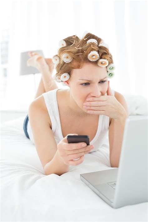 Shocked Woman In Hair Curlers With Cellphone Looks At Laptop In Bed