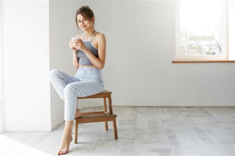 Young Tender Brunette Woman Smiling Holding Cup Sitting On Chair Over White Wall Early In