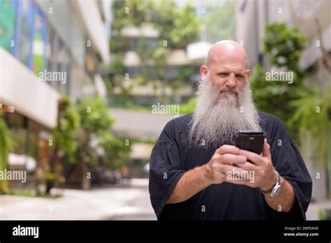 Mature Handsome Bald Bearded Man Using Phone With Nature In The City