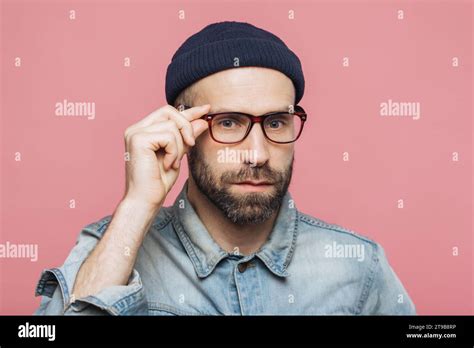 Serious Man Adjusting Glasses Wearing Beanie And Denim Shirt Against