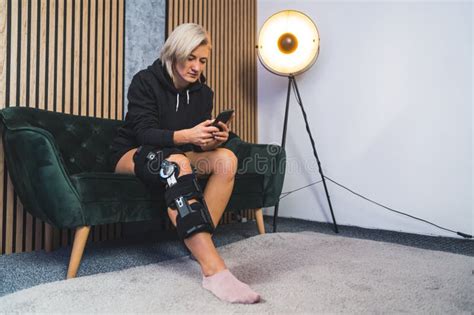 Woman With A Knee Brace Sitting On The Sofa And Holding Her Phone Stock