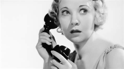 A Black And White Photo Of A Woman Holding An Old Phone To Her Ear While Looking At The Camera