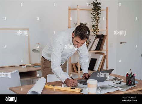 Mature Architect With Blueprint And Laptop Working At Desk Stock Photo