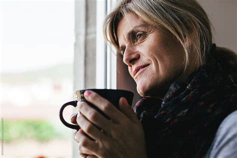 Mature Woman Holding A Cup Of Coffee Standing Next To The Window At Home By Stocksy