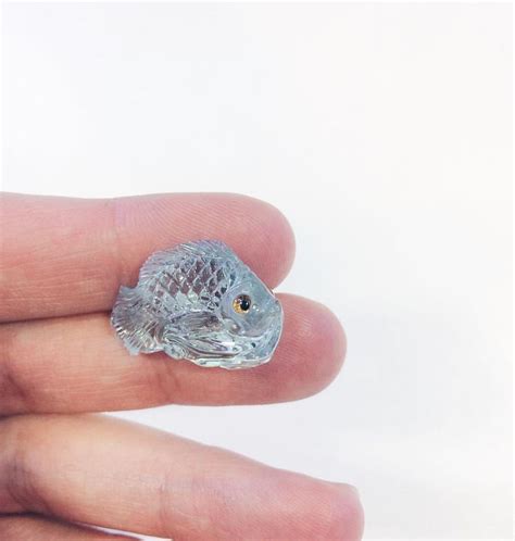 A Small Glass Fish Sitting On Top Of Someone S Finger In Front Of A