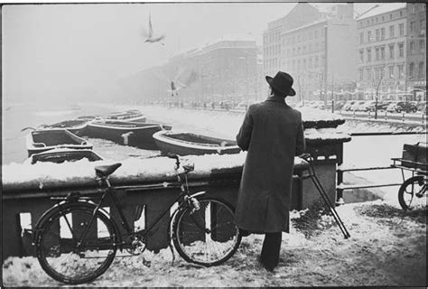 A Man Standing Next To Two Bikes In The Snow Near Some Boats And Birds