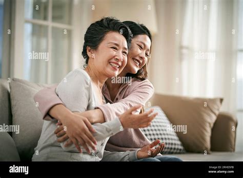 Loving Asian Adult Daughter Sitting On Couch At Home Hugging Senior