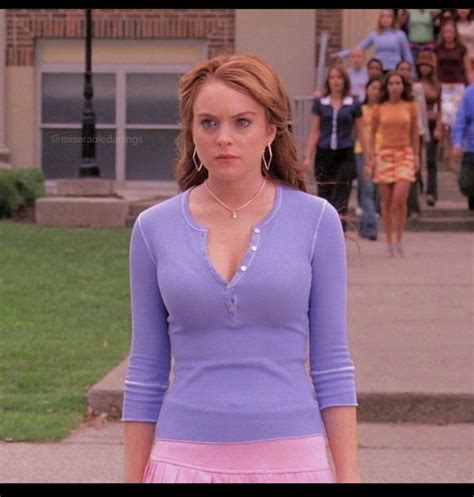 A Woman In A Purple Shirt And Pink Skirt Standing On The Sidewalk With People Walking Behind Her