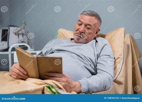 A Gray Haired Old Man Reading A Book In His Bed In A Hospital Ward