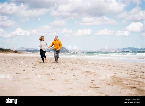 Cheerful Mature Couple Holding Hands Running On Shore At Beach Stock