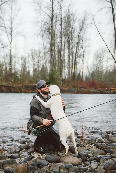 Fly Fisherman Receiving A Hug From His Puppy On The Bank Of A River