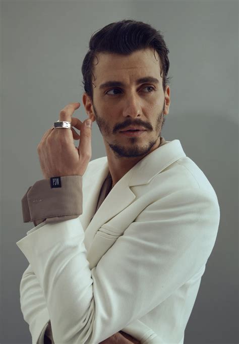a man in a white suit holding his hand on his ear and looking at the camera