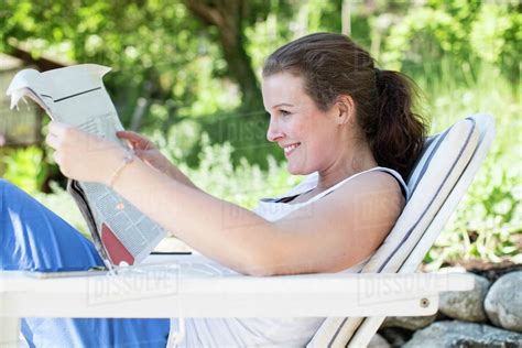 Side View Of Smiling Woman Reading Newspaper While Relaxing On Lounge