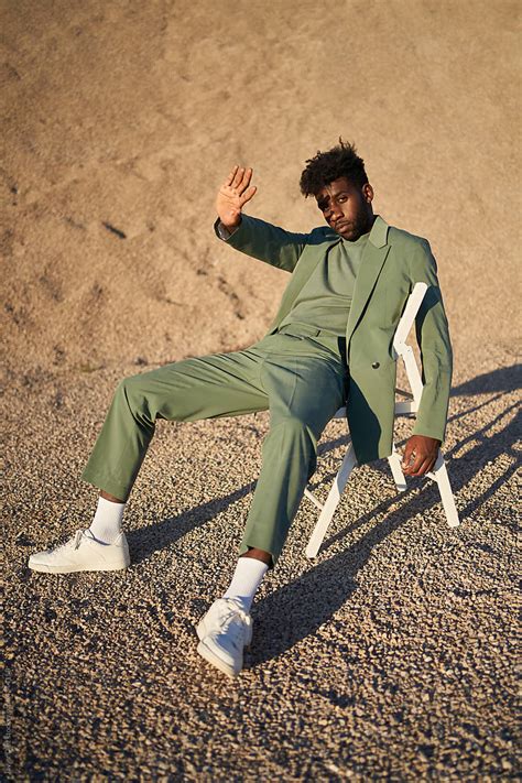 Stylish Black Man Sitting On Chair Against Sandy Hill By Stocksy