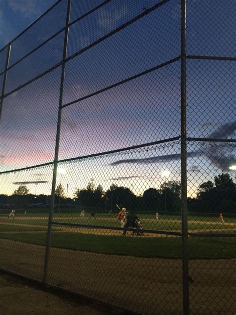 A Baseball Game Is Being Played Behind A Chain Link Fence At Dusk With