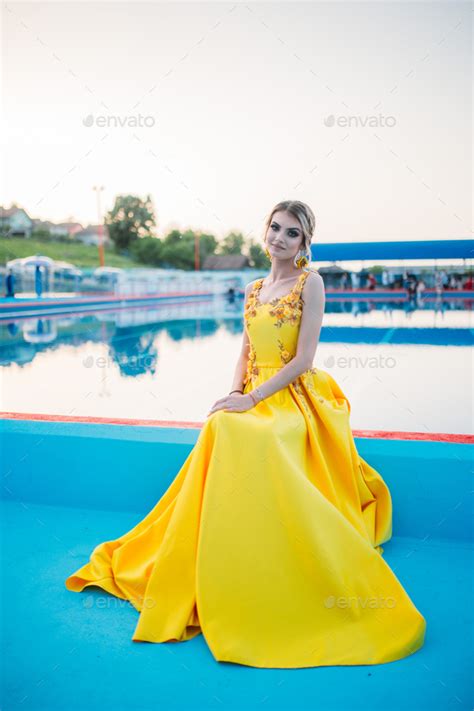 Gorgeous Blonde Female In An Elegant Yellow Dress Sitting Next To An Open Swimming Pool Stock