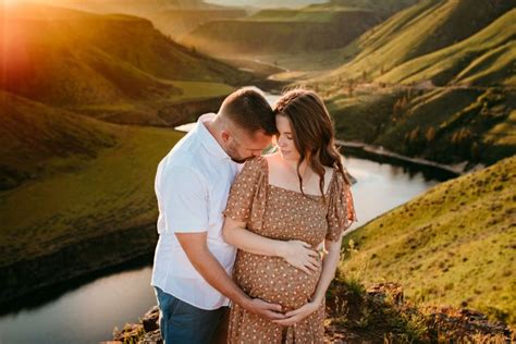 A Pregnant Couple Cuddles While Standing On A Hill Overlooking A River