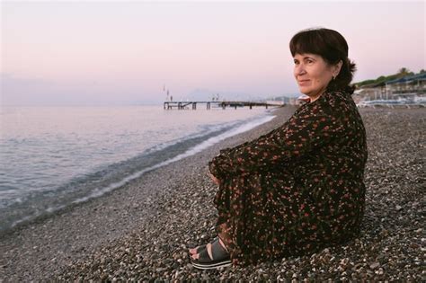Premium Photo Elderly Brunette Woman Sits At The Edge Of The Sea And Looks At The Sunset