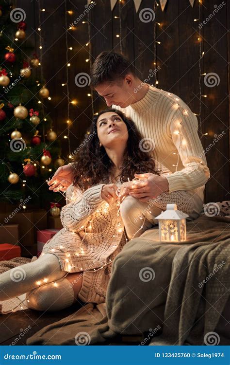 Young Couple Is In Christmas Lights And Decoration Dressed In White Fir Tree On Dark Wooden