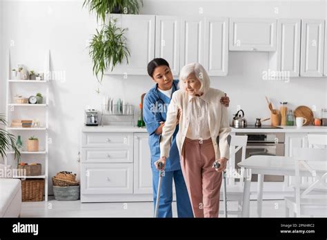 Brunette Multiracial Caregiver In Blue Uniform Helping Retired Woman