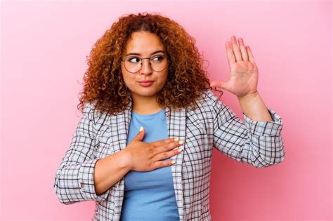 Joven mujer curvilínea latina aislada sobre fondo rosa tomando un juramento poniendo la mano en